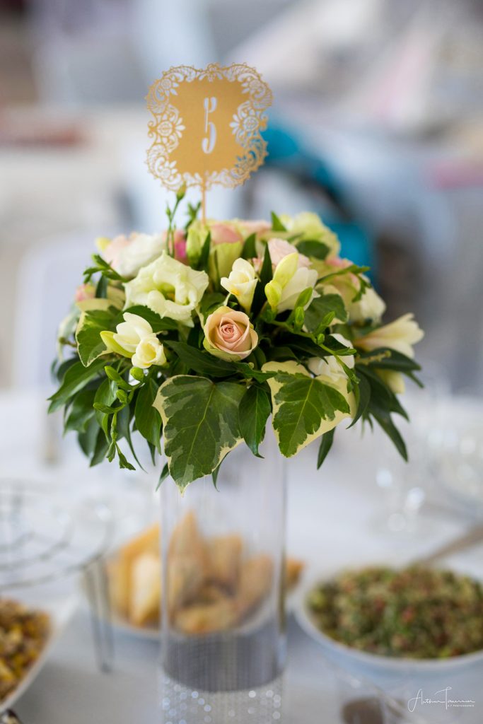 Composition florale de table lors d’un mariage, avec fleurs blanches et feuillage, photographiée dans un décor de réception.